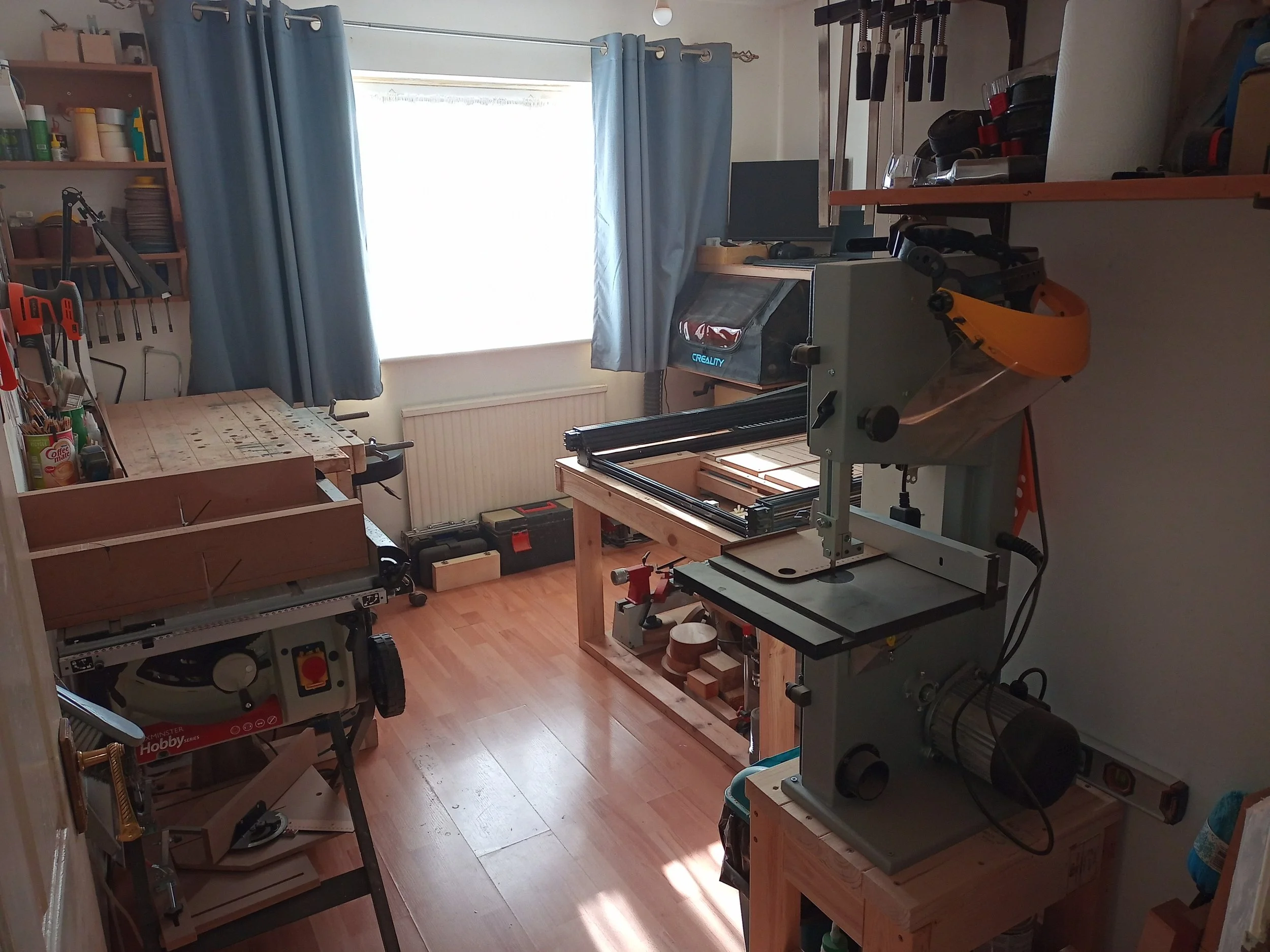 A woodworking workshop with a bandsaw, a tablesaw and a workbench. Also, there's a front edge of a CNC machine with a small lathe under it and a computer setup in the back corner sitting on top of a laser.