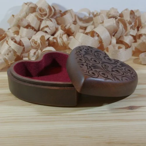 A heart-shaped jewelry box with intricate swirl designs on the lid, open to reveal a red interior, placed on a wooden surface with wood shavings in the background.