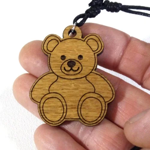A wooden teddy bear pendant with a black cord, held between fingers.