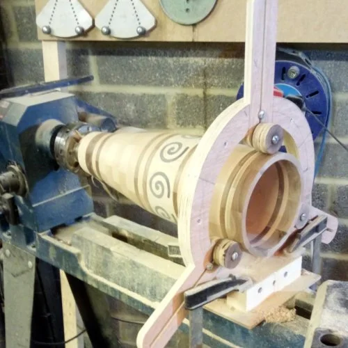 A wood lathe machine shaping a wooden cone with spiral patterns, in a workshop setting with tools and gauges on the wall.