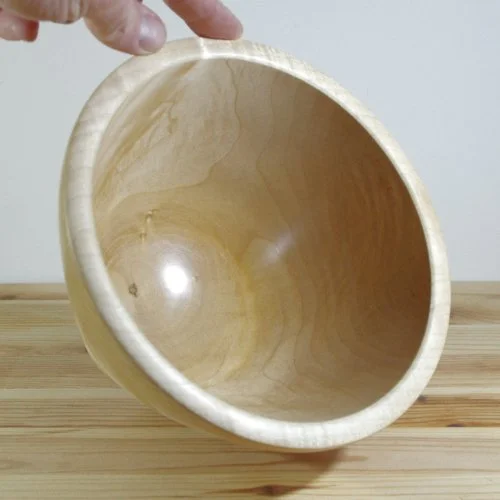 A hand holding a light-colored wooden bowl over a wooden surface with a plain background.