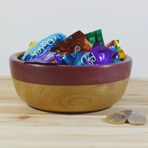 Cherry and purple heart bowl filled with assorted wrapped candies placed on a light wooden surface with a few coins nearby.