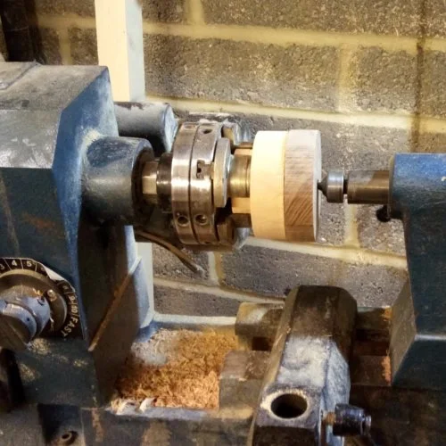 A woodturning lathe with a spinning wooden cylinder and metal components, mounted on a workbench in a workshop.