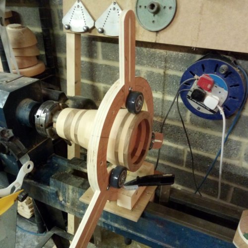 Woodworking lathe with a partially turned wooden piece mounted, in a workshop with tools and spools on the wall.