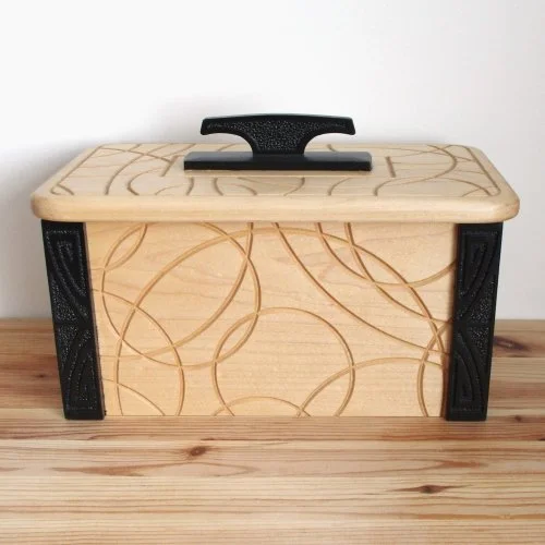 Wooden keepsake box with a lid featuring a circular pattern, black handle, and black decorative accents on the sides, placed on a wooden surface.