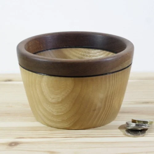 A wooden bowl with a darker rim on a light wooden surface, with a small stack of coins beside it.