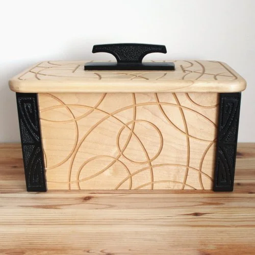 A wooden keepsake box with black handle, featuring engraved swirling line patterns on its surface, placed on a wooden table against a plain white wall.