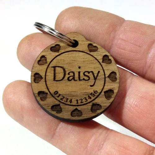 Wooden pet tag with the name 'Daisy' engraved, surrounded by small heart shapes, and a phone number engraved at the bottom.