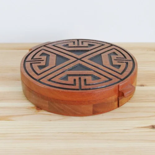 Round jewellery box with intricate black geometric pattern on the top, placed on a light wooden surface.