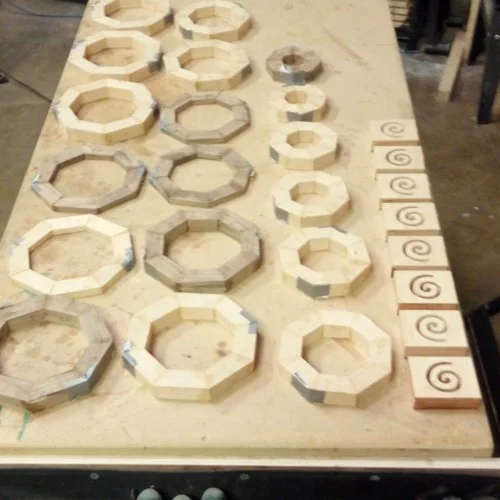 Wooden octagon-shaped and smaller square frames with spiral carved designs on rectangular blocks on a workbench.