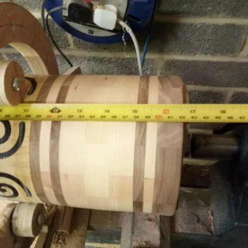 A partially turned wooden vase on a lathe with a yellow measuring tape across the diameter, in a woodworking workshop.