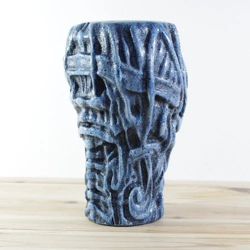 Blue wooden vase with a carved abstract skull design, placed on a light wooden surface.