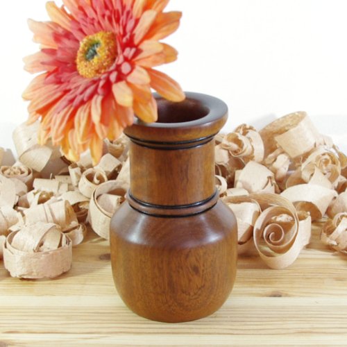 A wooden vase holding a pink and orange flower with a yellow center, surrounded by wood shavings on a wooden surface.