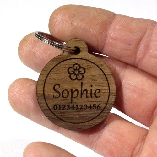 Wooden circular pet tag with a flower design, the name Sophie, and the numbers 01234123456, held by a person's fingers