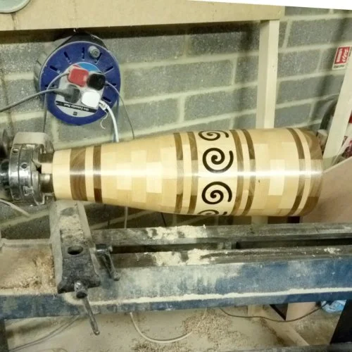 Woodworking lathe machine turning a wooden piece with decorative spiral and geometric patterns.