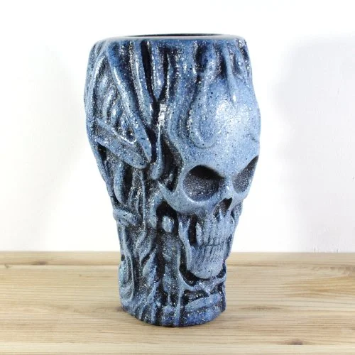 A wooden vase with a textured, blue and black skull design, placed on a wooden surface against a plain white background.
