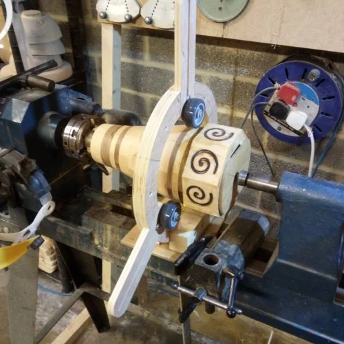 Wooden cylindrical object with spiral and swirl patterns mounted on a lathe in a workshop.