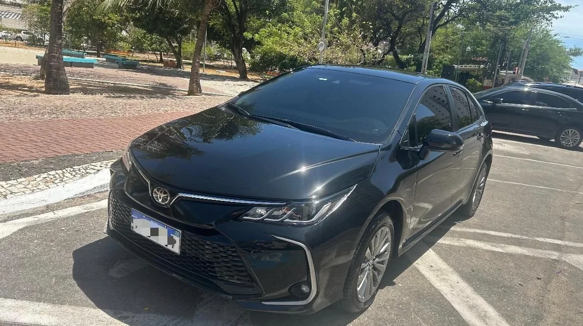 Carro preto estacionado em uma área de estacionamento ao ar livre, com árvores ao fundo.