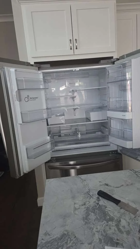Open empty stainless steel refrigerator in a modern kitchen with white cabinets and granite countertop.