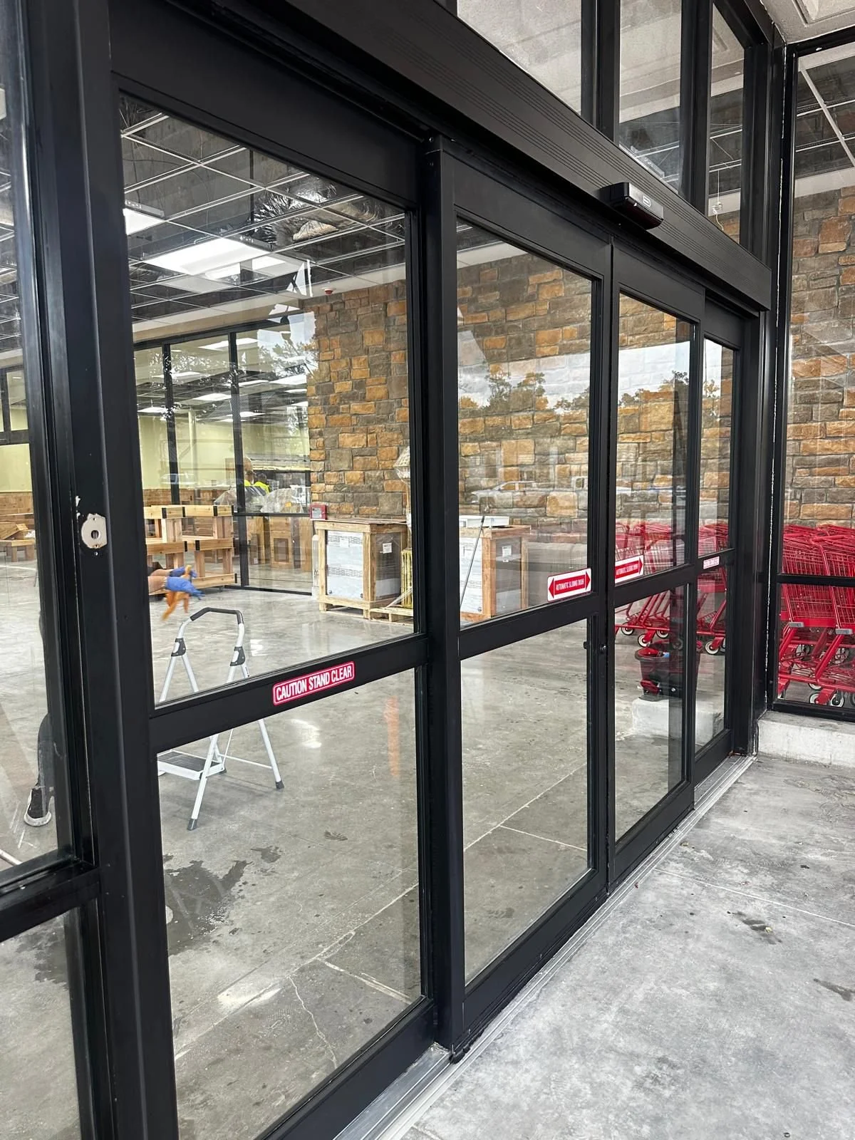 Glass sliding door at the entrance of a store or building with a brick wall interior, red shopping carts stacked to the side, and a caution sign on the door.