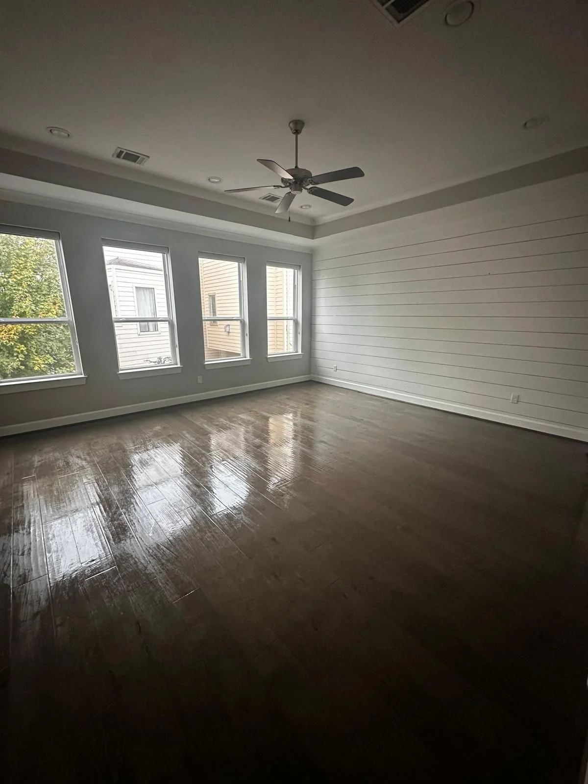 Empty living room with hardwood floors, white walls, four windows, a ceiling fan, and natural light coming in.