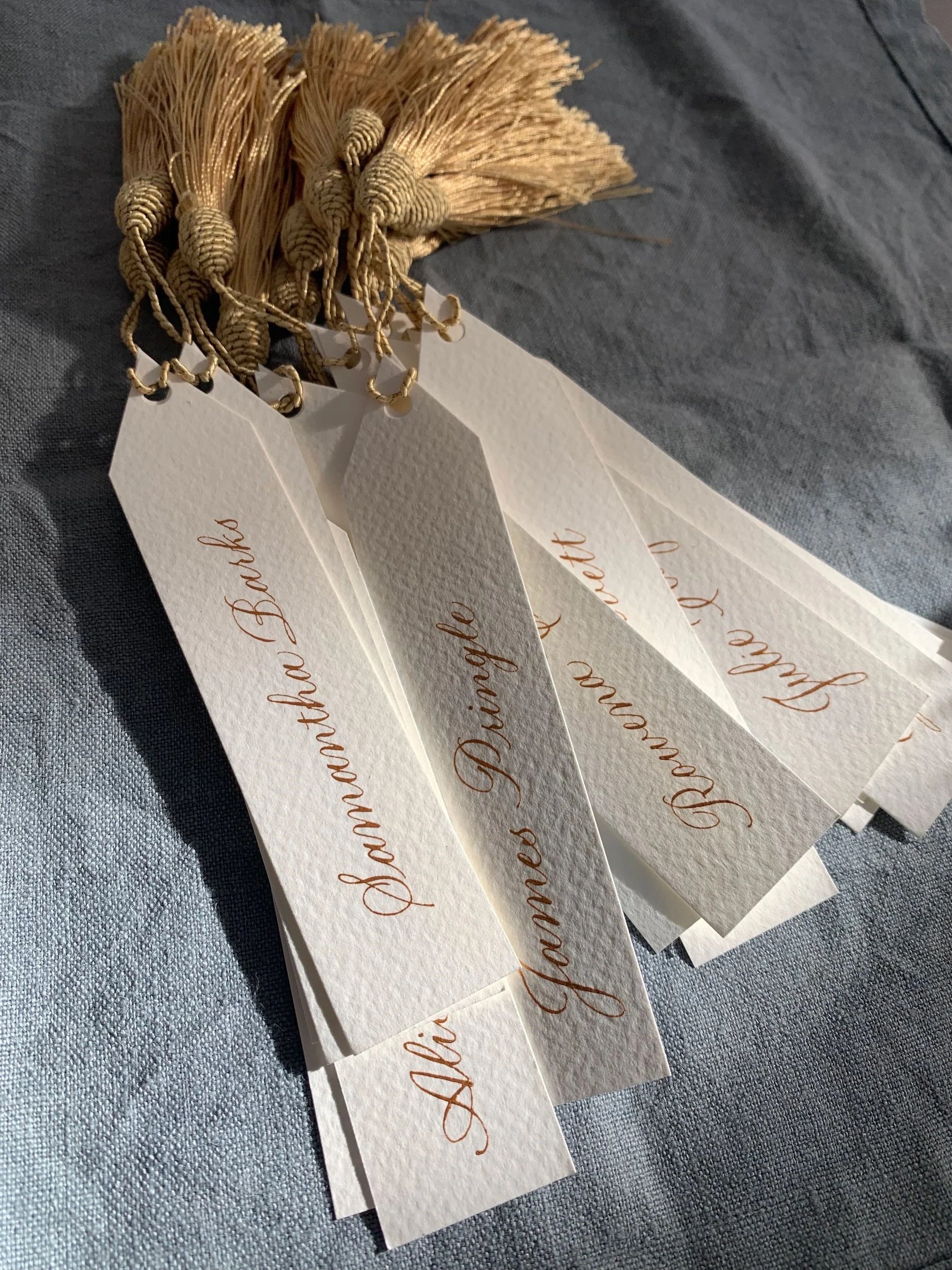 Several wedding program or ceremony cards with gold calligraphy on white textured paper, attached to small straw fans, resting on a gray fabric surface.