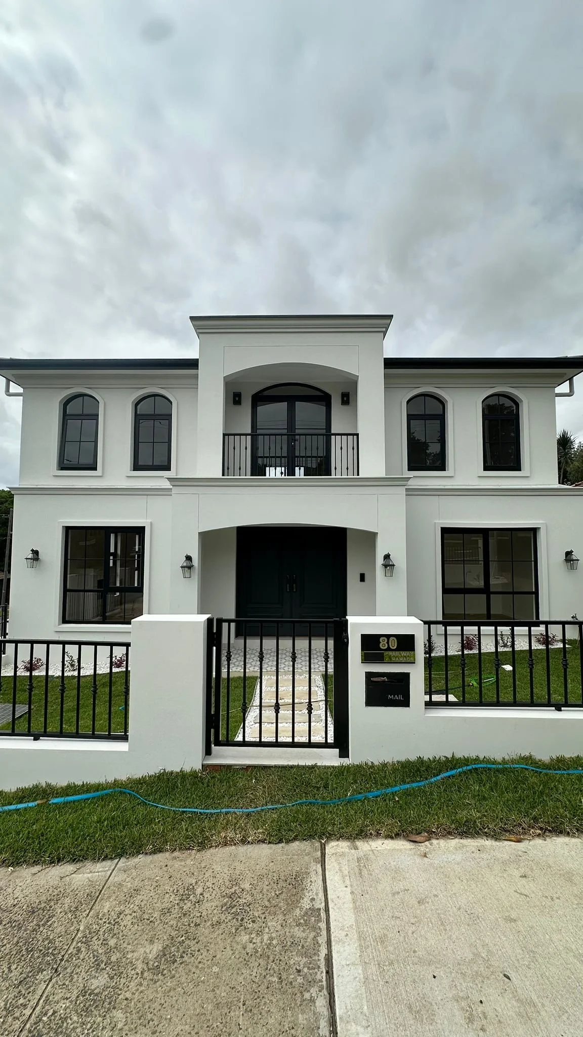A modern two-story white house with black window frames, a black front door, and a small front yard with a black fence. The house features arched windows and a balcony on the second floor. The sky is cloudy.