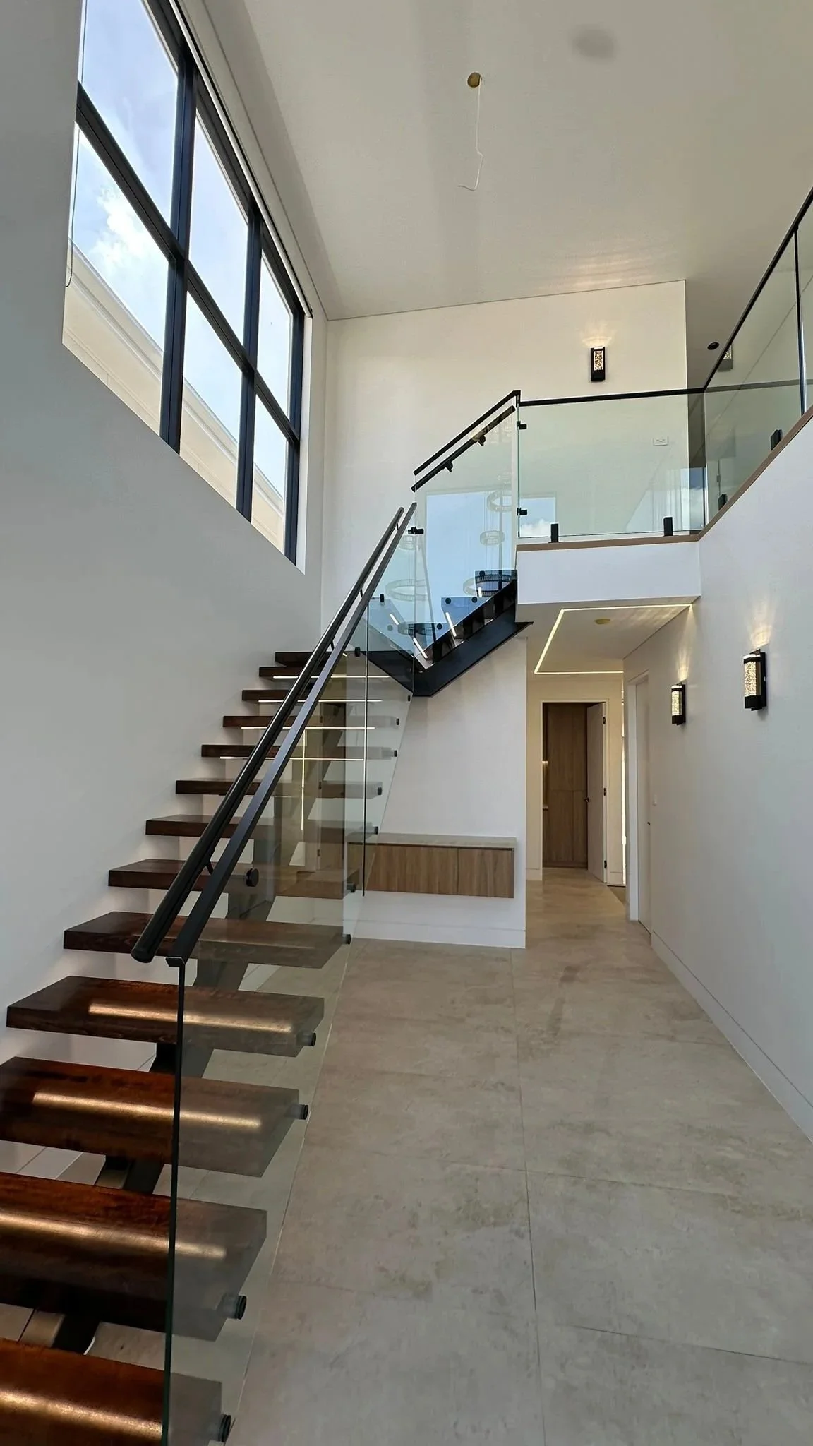 Modern staircase with wooden steps and glass railing inside a spacious house with large windows and minimalist decor.