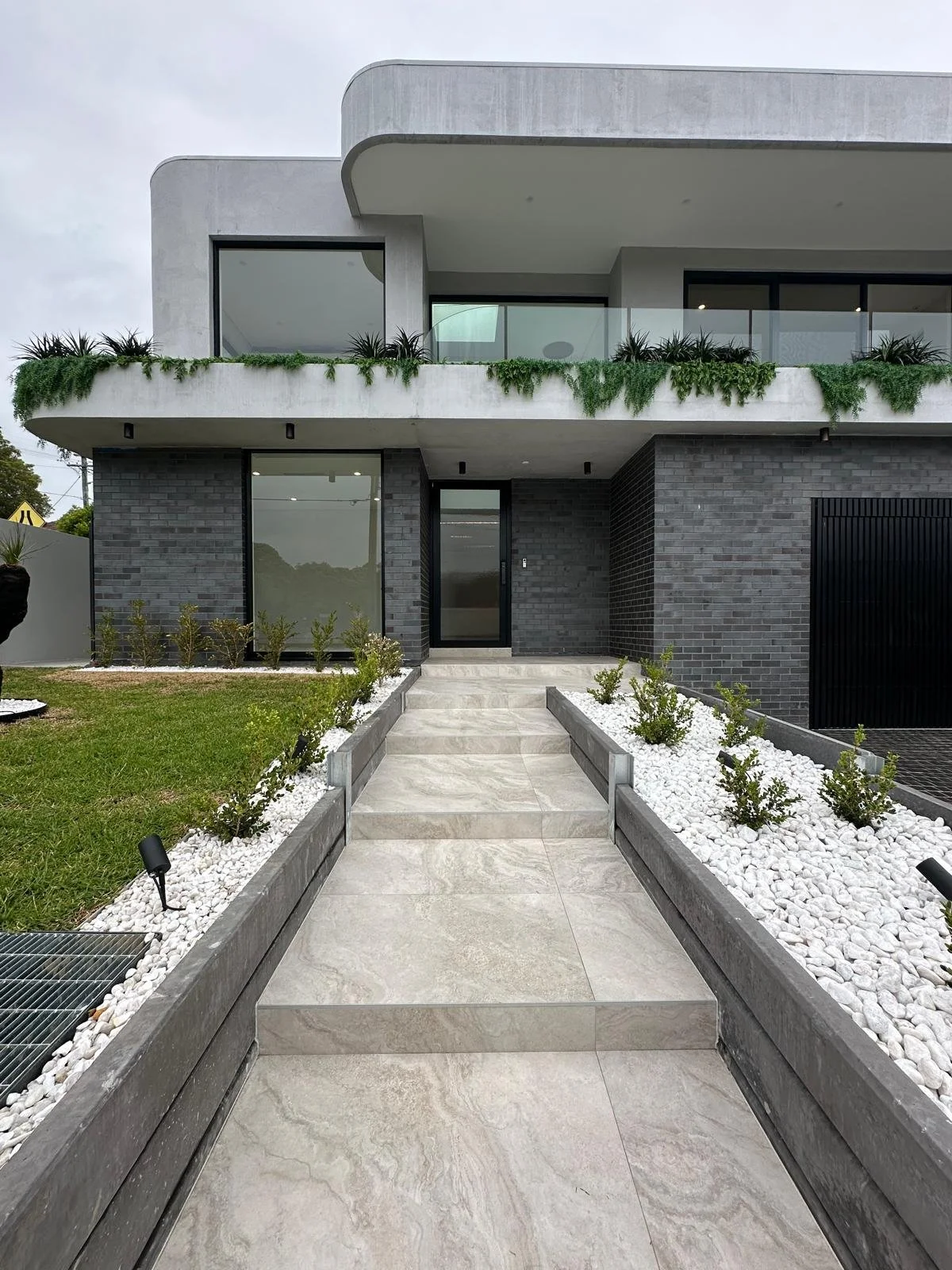 Modern gray house with a staircase leading to the front door, landscaped with white rocks, green plants, and potted plants on the balcony.