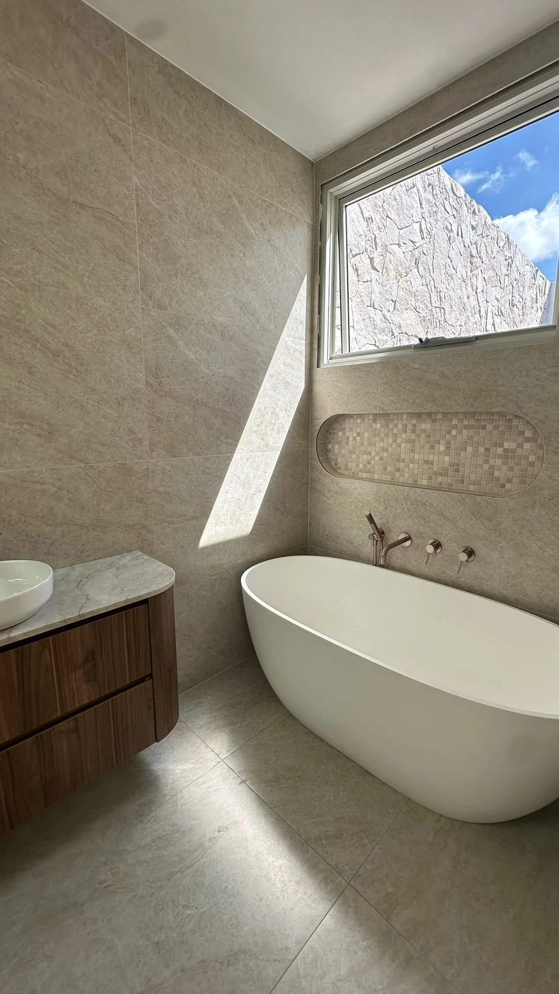 Modern bathroom with beige tiled walls and floor, a freestanding bathtub near a window with a view of the blue sky and a stone wall outside, and a wooden vanity with a marble top and a round white vessel sink.