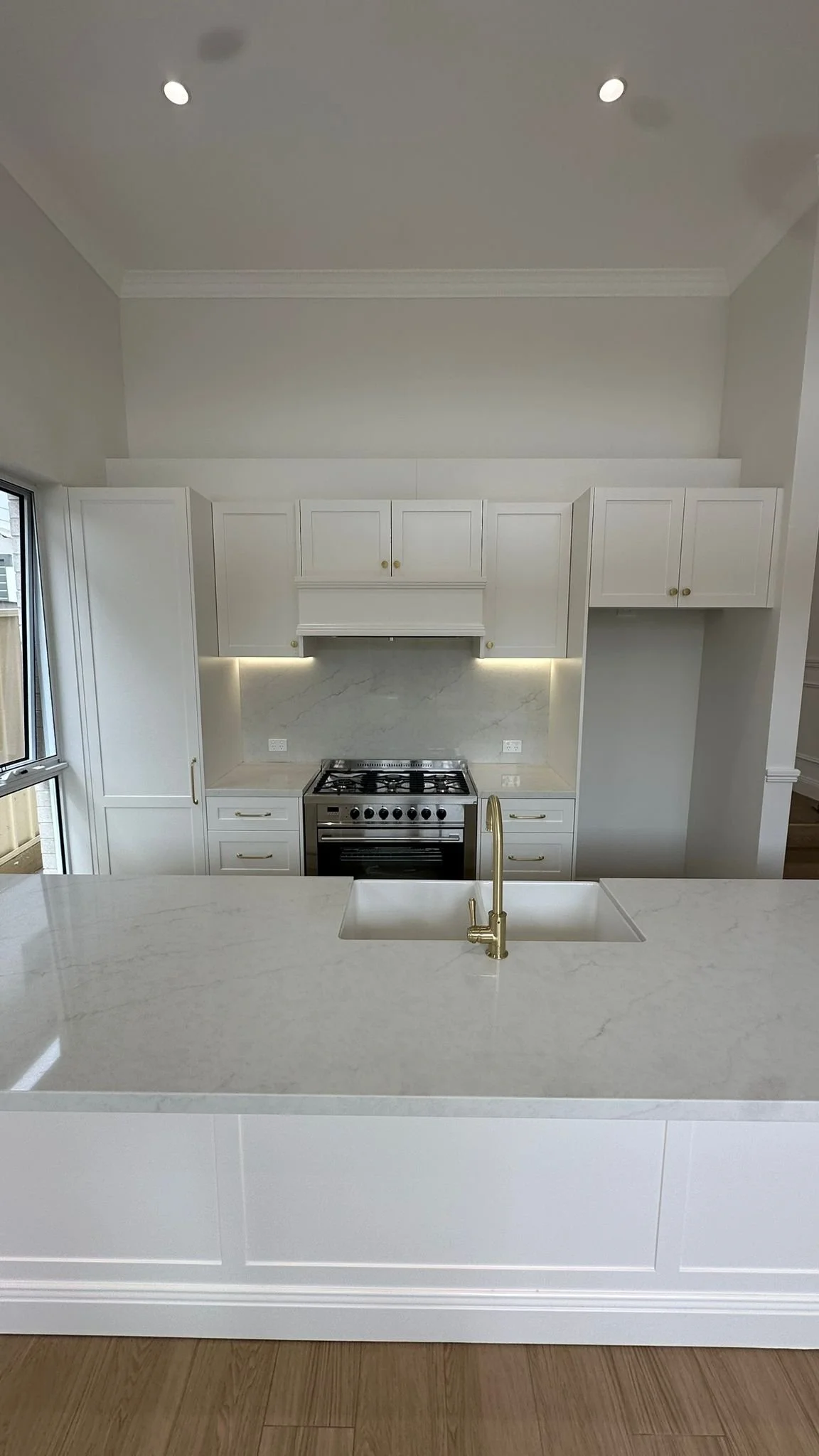Modern white kitchen with marble countertops, built-in stainless steel oven, gold faucet, and upper and lower cabinets with gold handles. A window on the left side allows natural light.