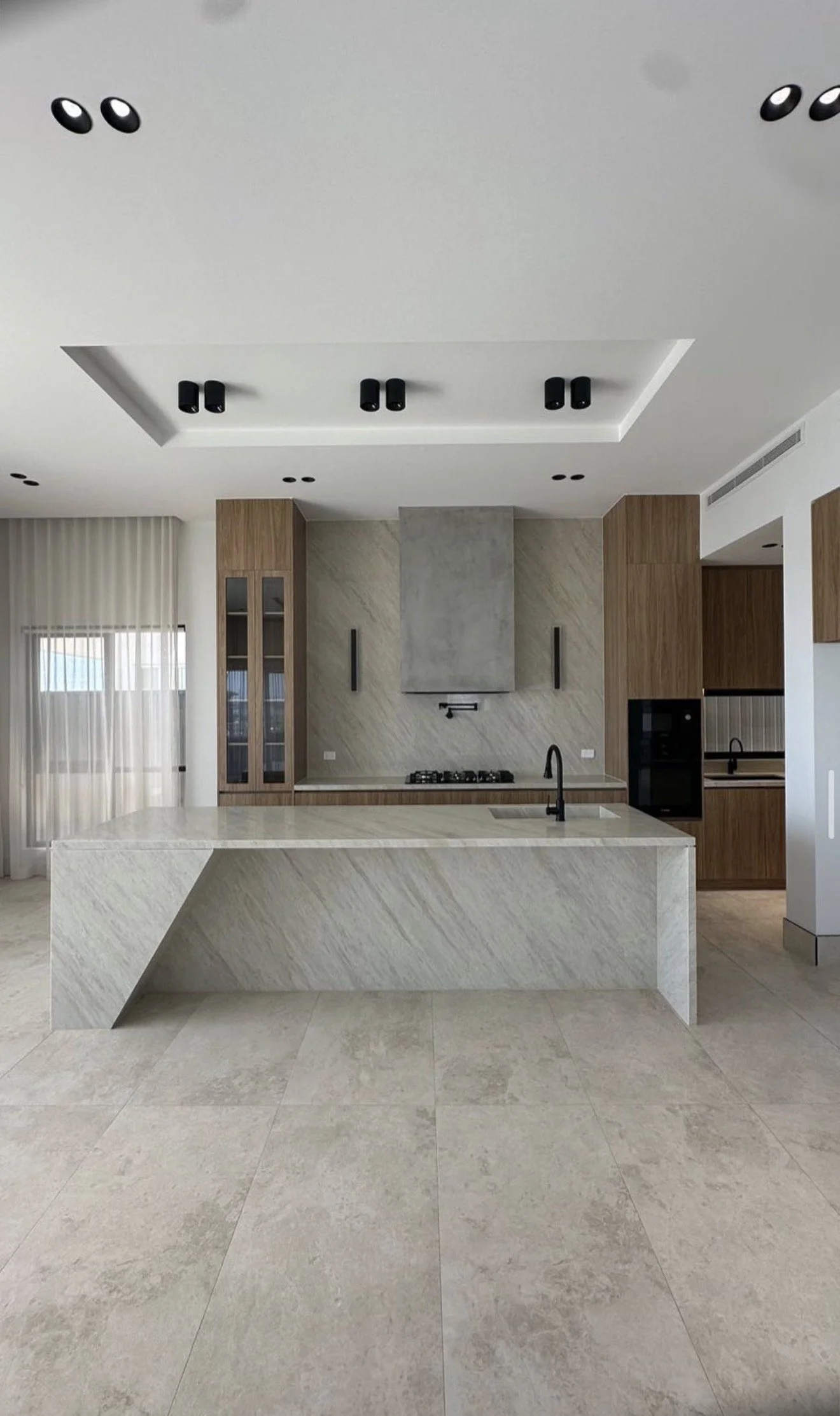 Modern kitchen with marble island, black faucet, built-in appliances, light-colored tiled floor, and wooden cabinetry.