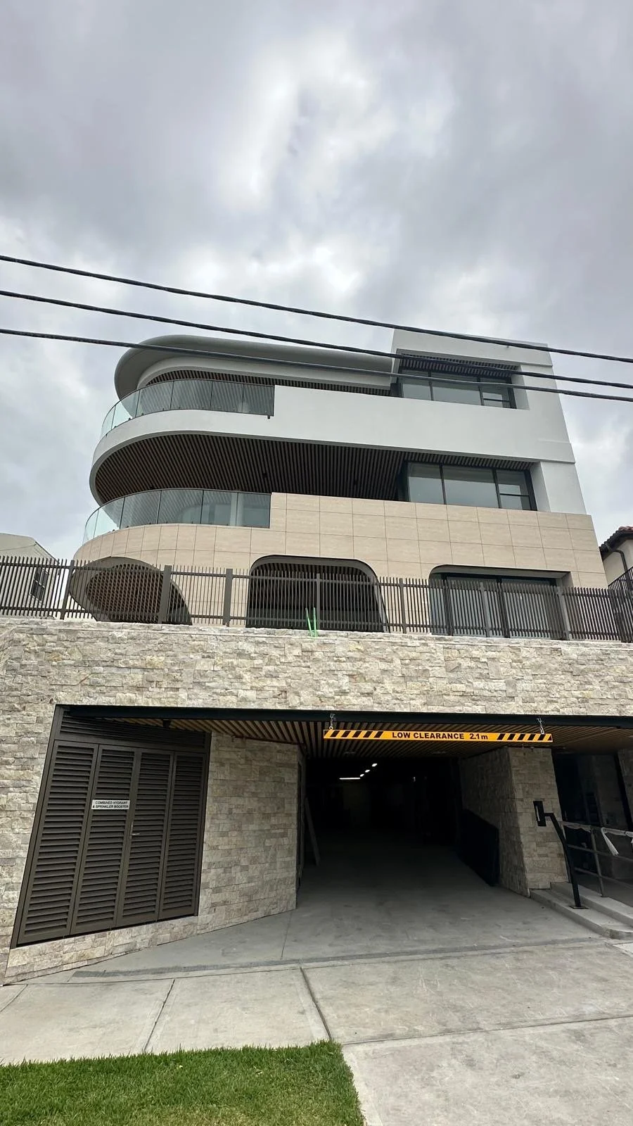 Modern multi-story building with curved balconies, glass railings, beige stone and gray exterior, and an entrance to a parking garage marked with a low clearance sign on a cloudy day.
