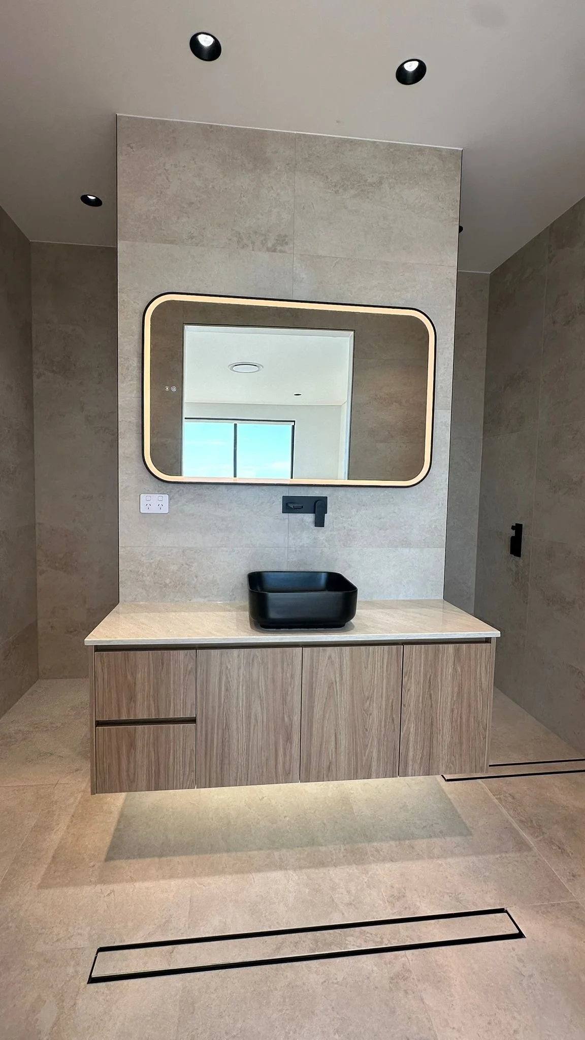 Modern bathroom with a rectangular mirror, wooden vanity, black sink, and black faucet, with a window reflected in the mirror and black recessed ceiling lights.