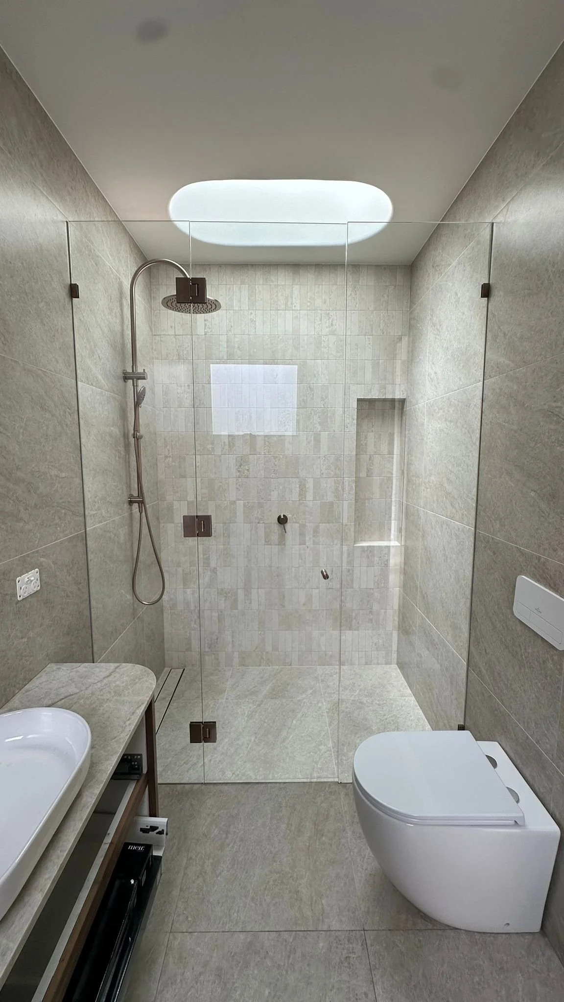 Modern bathroom with a walk-in shower with glass door, a toilet, and a sink with a countertop. The shower has a ceiling-mounted rainfall showerhead and a small built-in niche. The walls and floor are tiled in neutral tones, and there is a window in the shower area.