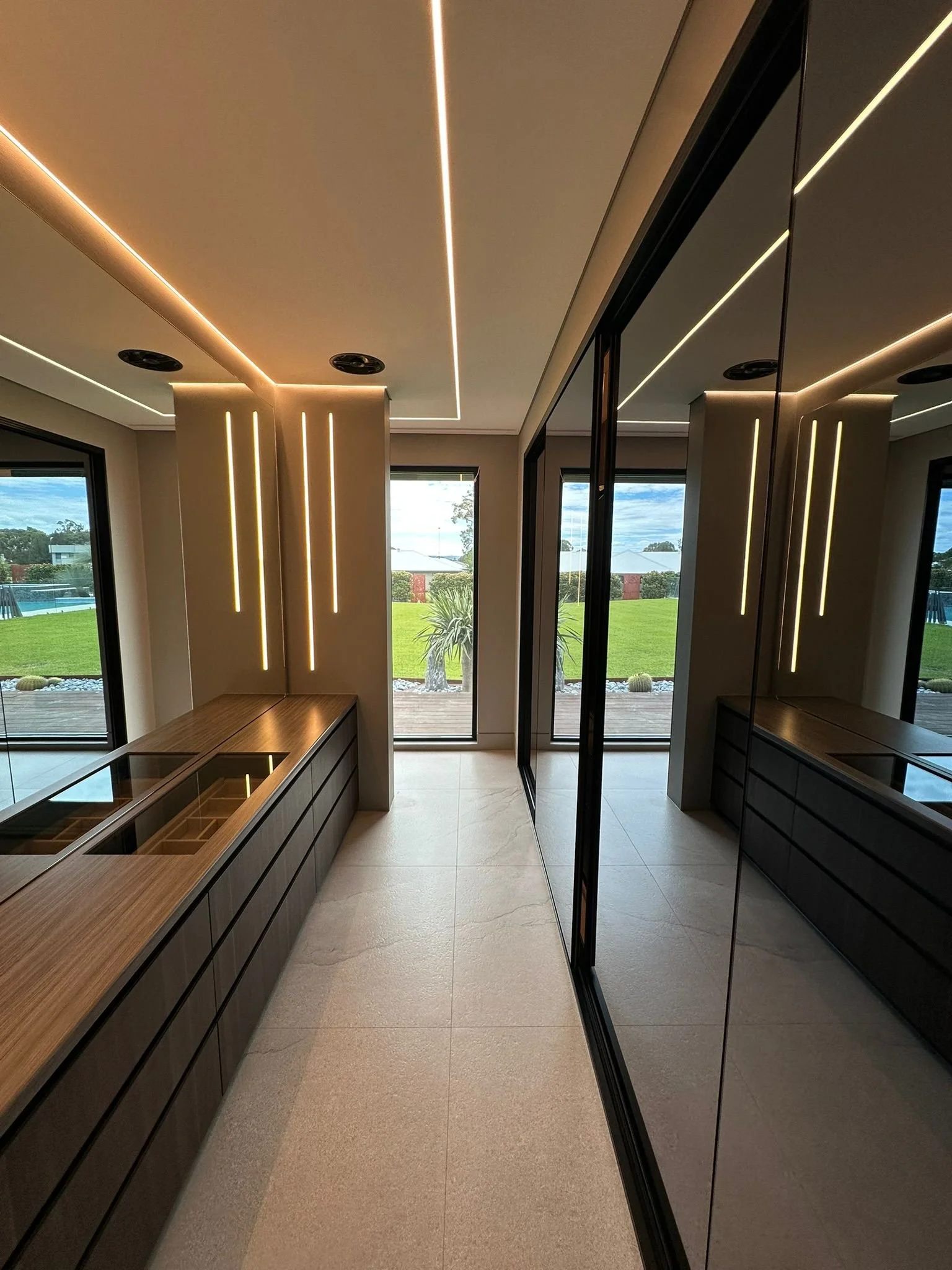 Modern kitchen with large windows, view of a grassy backyard, wooden cabinets with glass covers, mirrored closet doors, and LED strip lighting on the ceiling and wall.