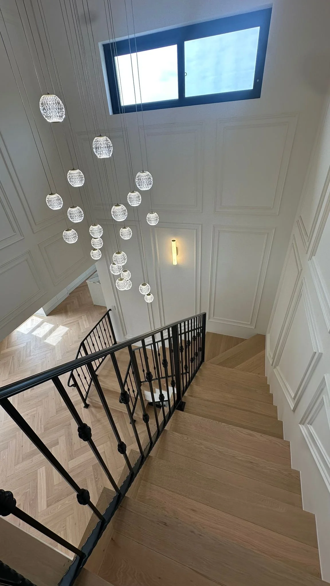 View of a staircase with wooden steps, black metal railing, modern hanging light fixtures, a wall-mounted light, and a large window in the ceiling showing the sky.