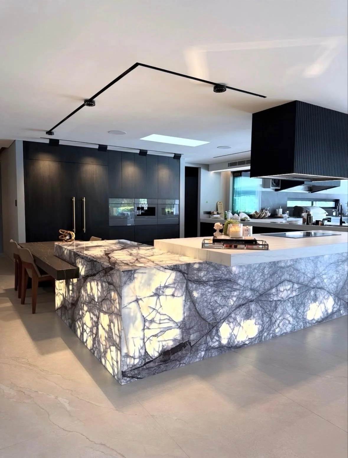 Modern kitchen with a large illuminated marble island, black cabinetry, exposed black ceiling light track, and a skylight in a contemporary home.