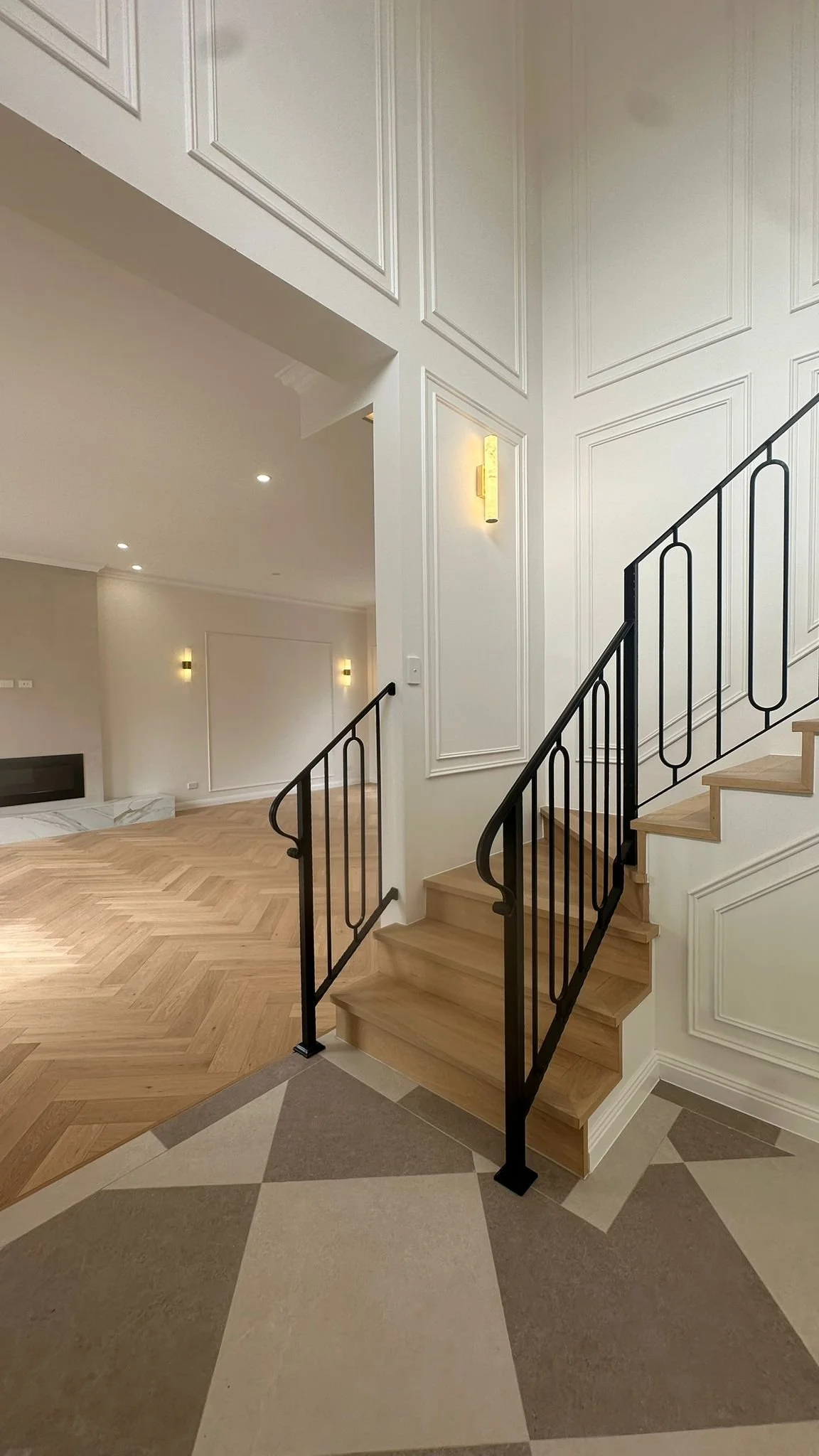 Interior of a modern home with a staircase, black metal railing, and a mix of tile and wooden flooring. White paneled walls and built-in lighting.