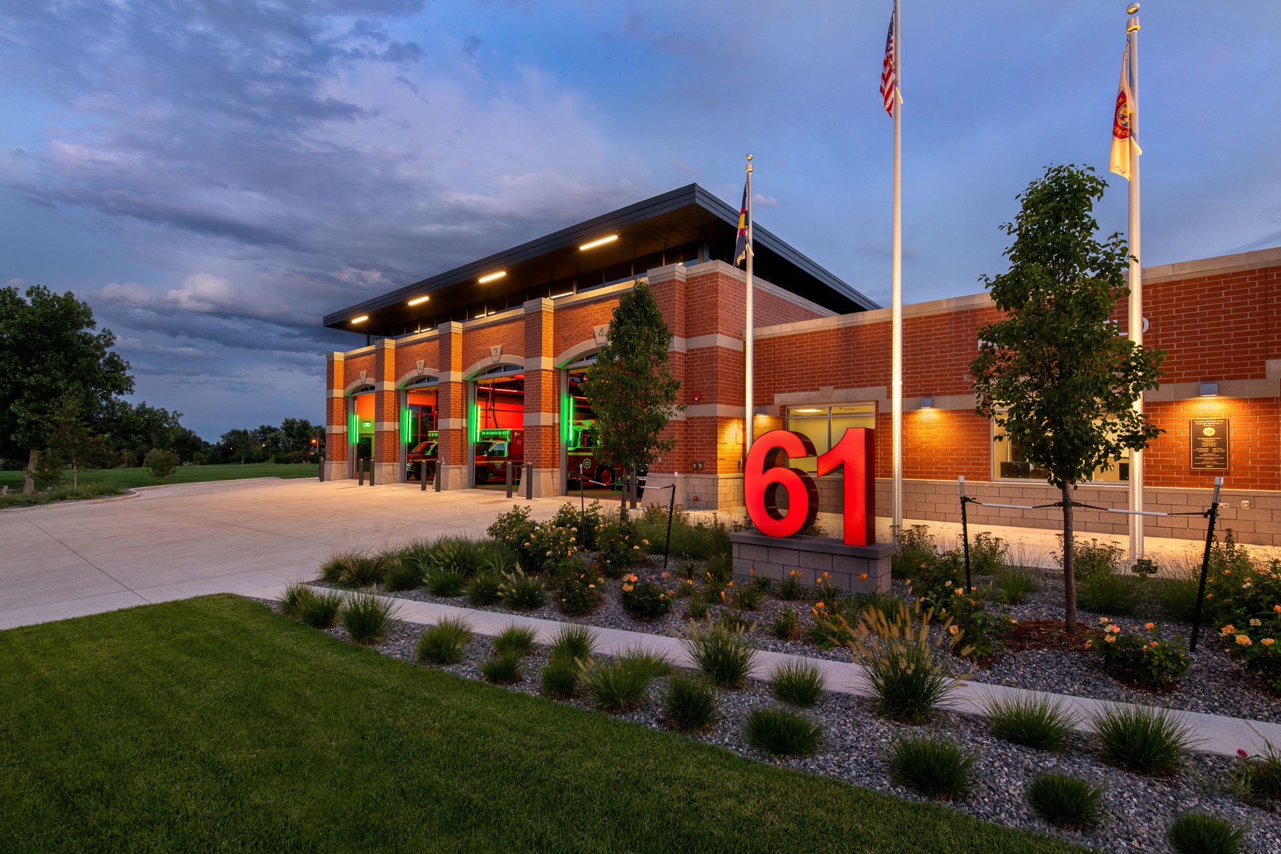 Dusk exterior of Fire Station 61, a brick building with illuminated truck bays, red trucks visible inside,  and landscaped entrance with red "61" signage.
