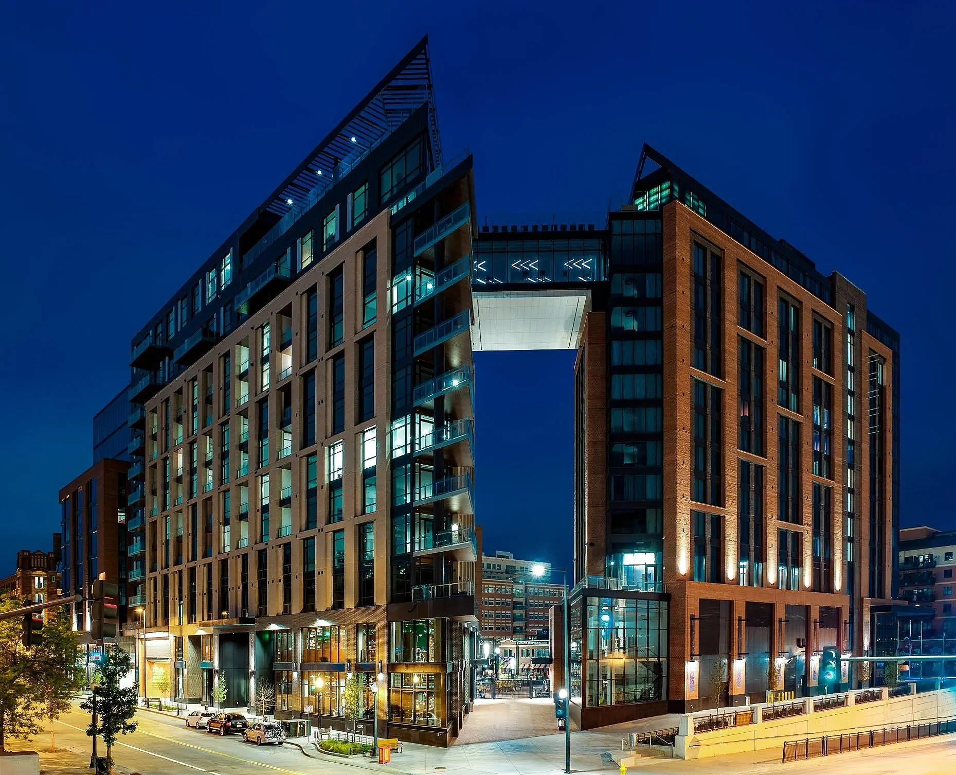 Twilight exterior of McGregor Square Residences, a multi-story mixed-use building with two illuminated wings connected by an elevated glass skybridge, ground-floor retail, and an urban street scene below.