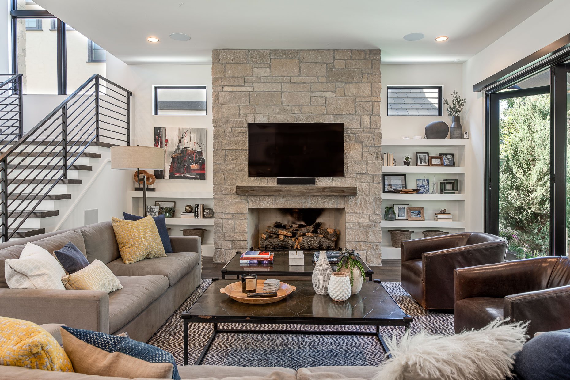 Cozy family room with couches and chairs, rock fireplace and open bookshelves flanking the fireplace. Nano doors open out to the patio.
