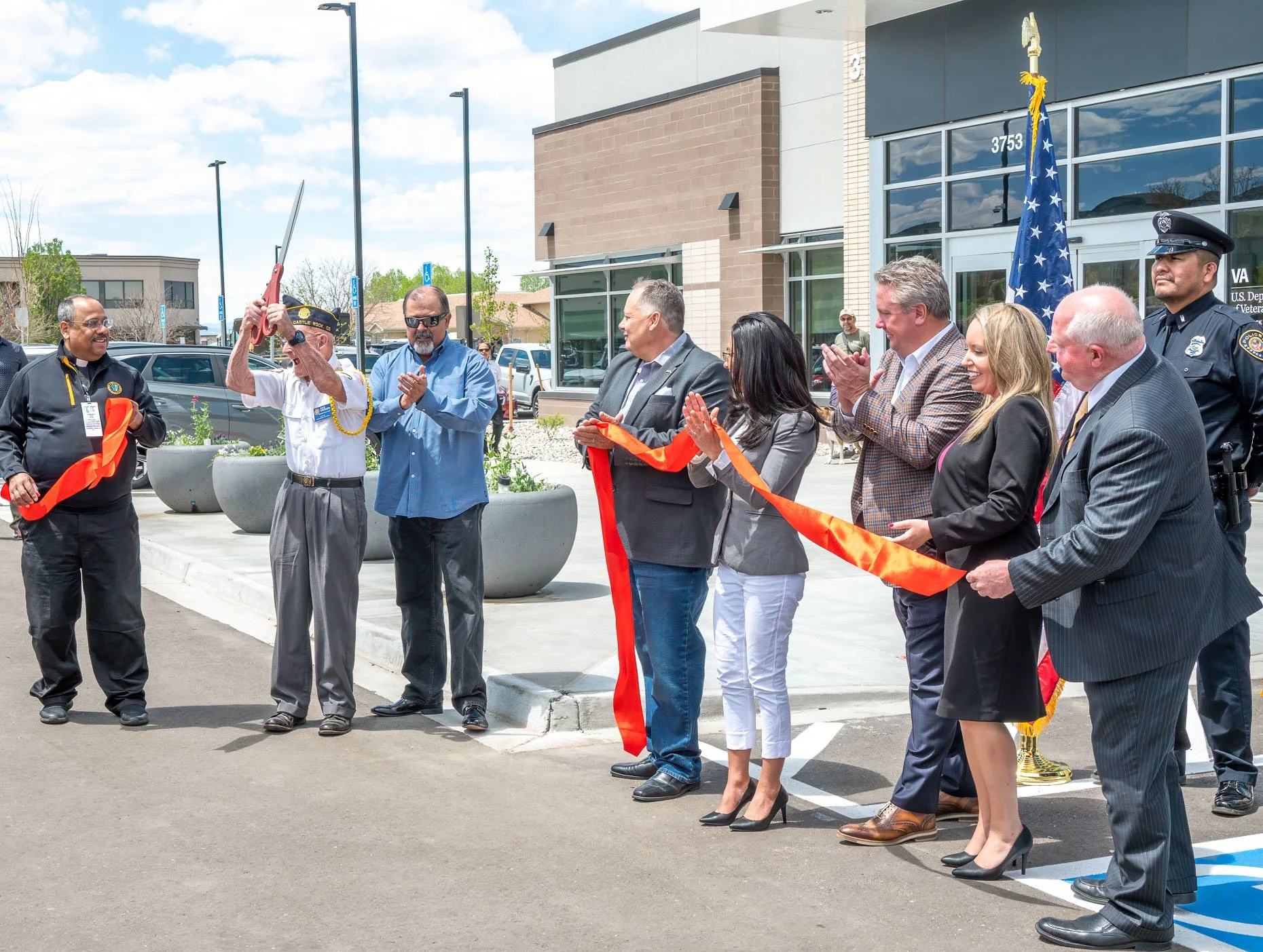 A group of people, including officials and a police officer, participating in a ribbon-cutting ceremony outside a new building, with some clapping and others holding a ribbon.