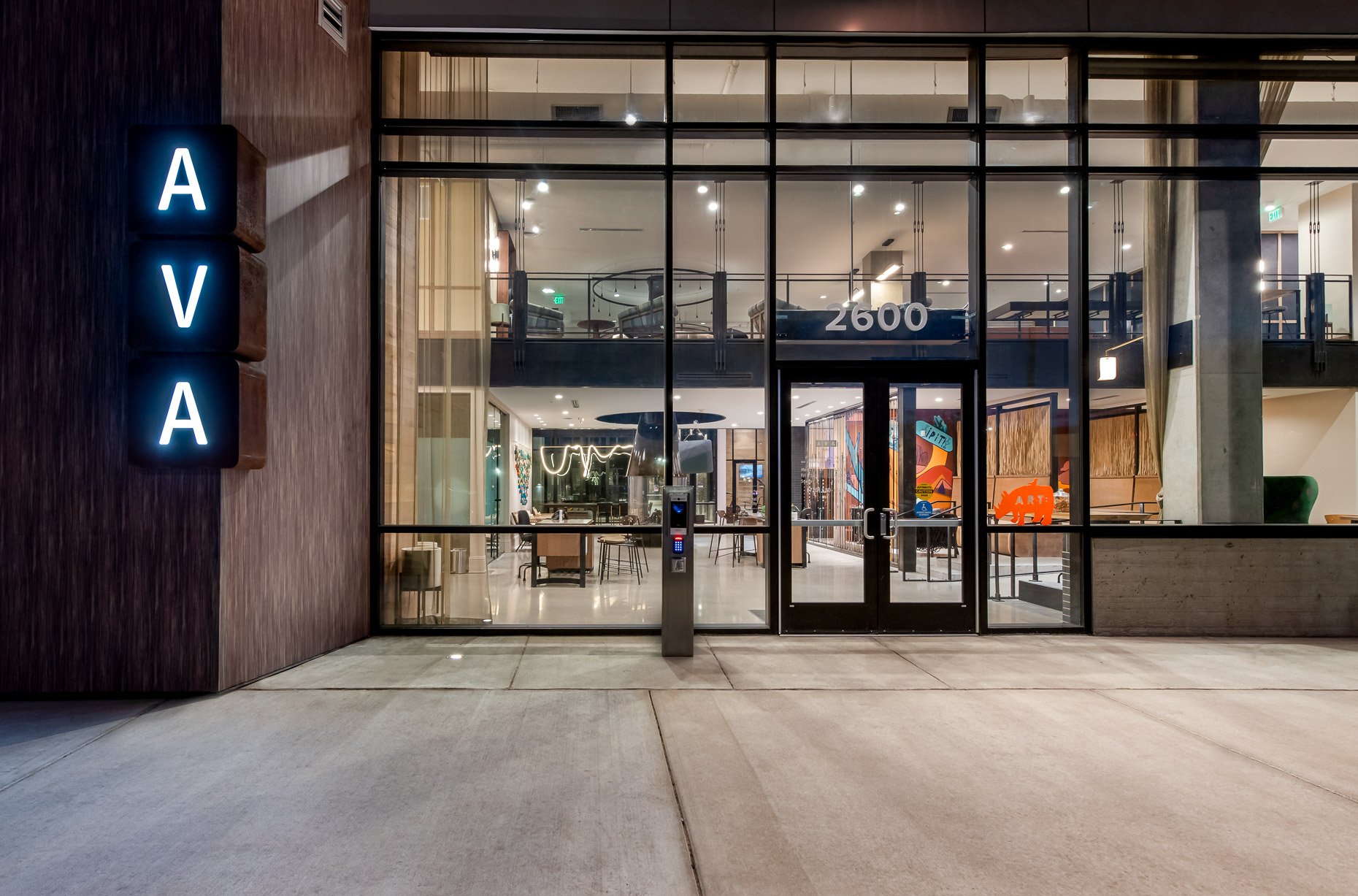 Exterior night view of AVA RiNo apartments at 2600, featuring floor-to-ceiling glass facades, illuminated lobby interior, and vertical neon signage.