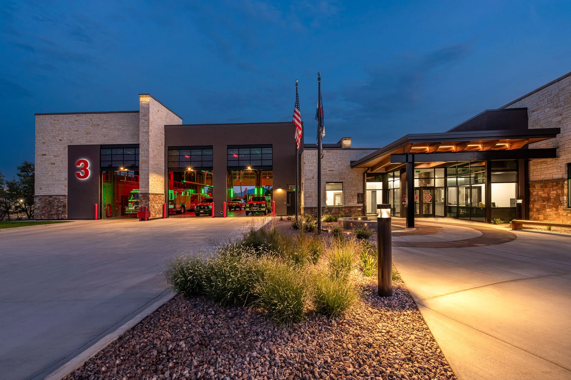 Twilight exterior of Fire Station 3 in Cañon City, a stone building with illuminated apparatus bays showing fire trucks, an entrance canopy, flagpoles, and landscaped beds along the driveway.