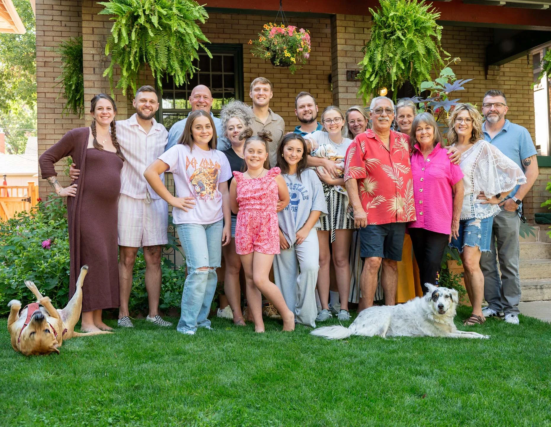 A large group of people, including children, adults, and seniors, gathered outdoors in front of a brick house with hanging plants and greenery. They are smiling and posing for the photo, with a few dogs sitting on the grass in front of them.