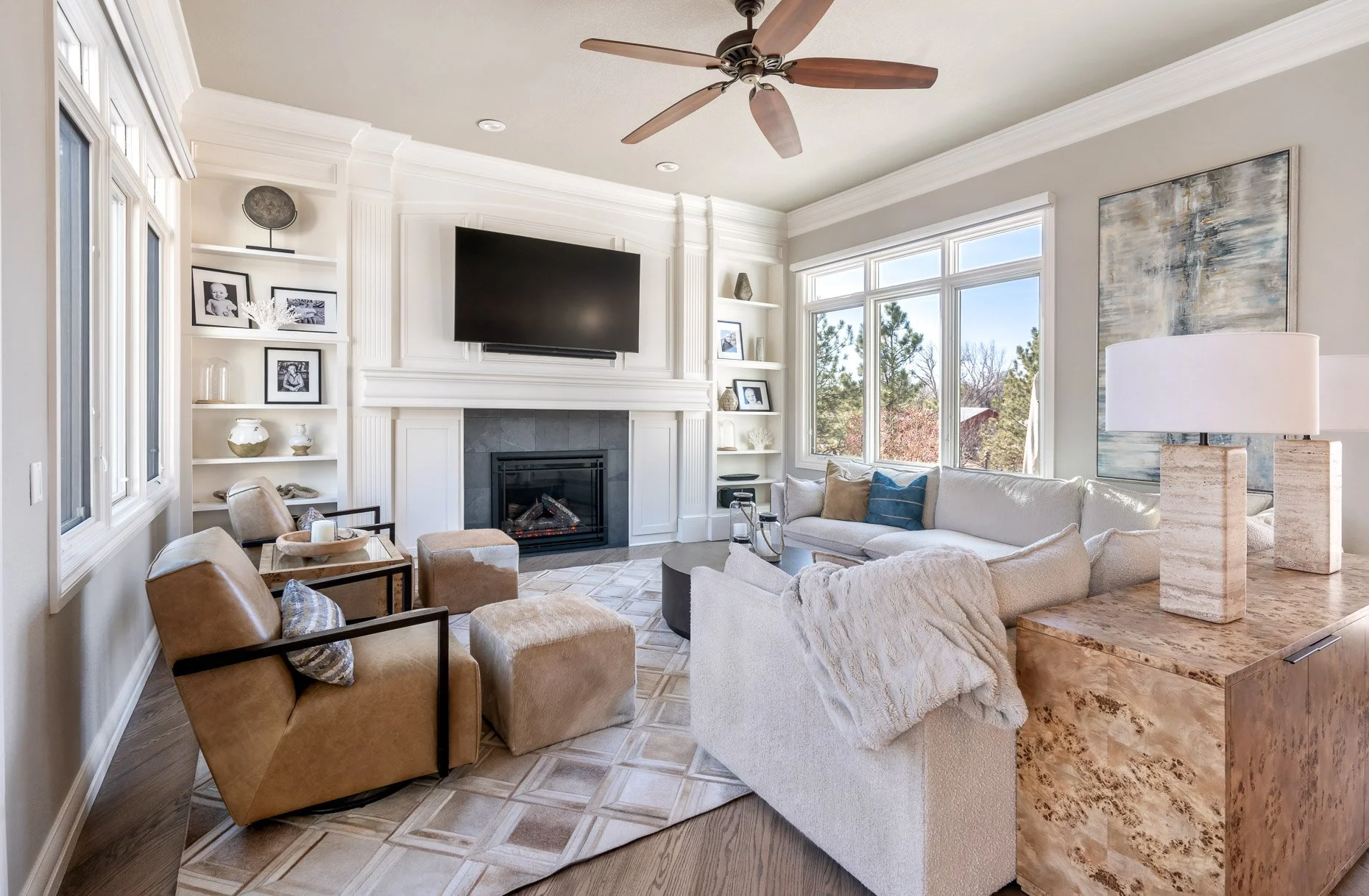 Living room with white built-in shelving and fireplace surround, mounted TV, leather swivel chairs, a cream sectional sofa with throw blanket, patterned area rug, and a burl wood console table with travertine lamps.