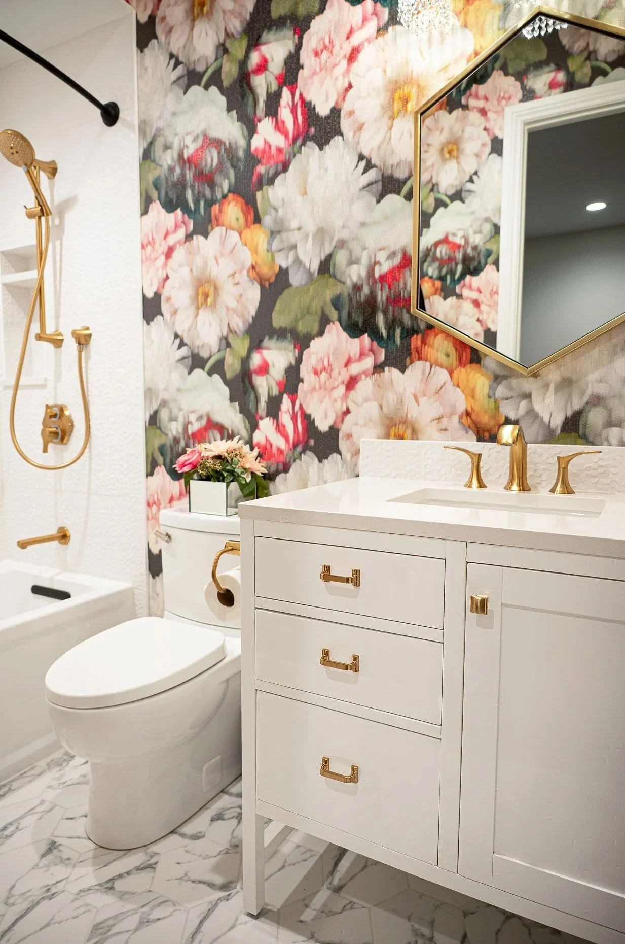 Bathroom with bold dark floral wallpaper, white vanity with brass hardware and fixtures, hexagonal brass-framed mirror, marble floor tile, and a tub/shower with gold fixtures.