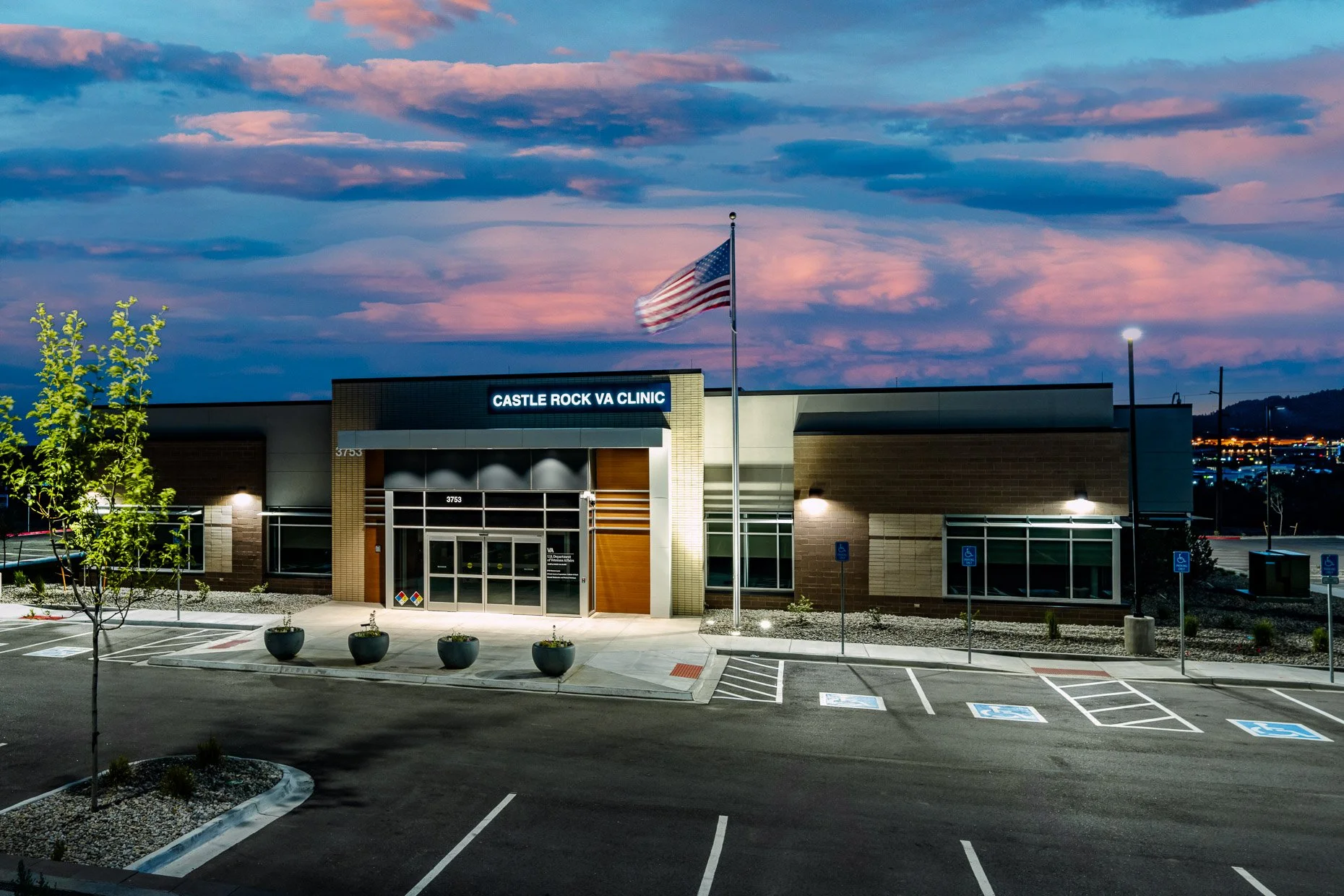 Exterior of the Castle Rock VA Clinic at dusk, with an American flag flying in front of the modern brick and stone building, illuminated against a dramatic pink and blue sunset sky.