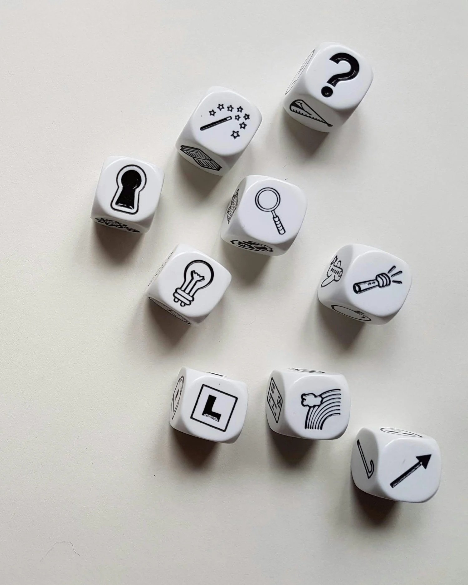 Set of nine white dice with black icons and symbols, including a keyhole, star gauge, question mark, book, magnifying glass, light bulb, rainbow, arrow, and ladder, scattered on a white surface.